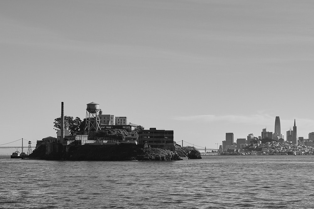 Black and white landscape photo of Alcatraz Island with its iconic water tower in the foreground. The island is contrasted by the city of San Francisco in the background.