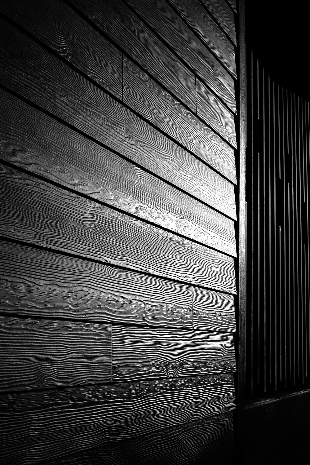 Black and white portrait photo of a side of a building clad in wood. The mid-day sun shows the detail in the grain of wood creating interesting textures.
