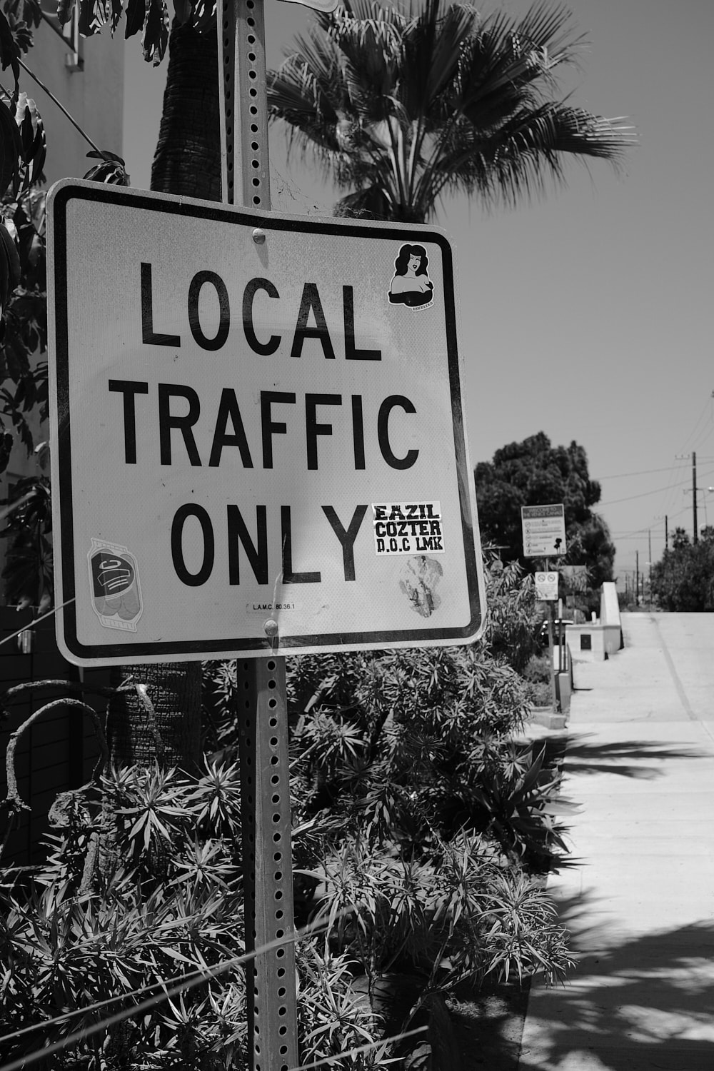 Black and white portrait photo of a street side just before one of the Venice canal bridges. The sign reads LOCAL TRAFFIC ONLY in all caps. The sign is covered with stickers and is all bent and beaten up.