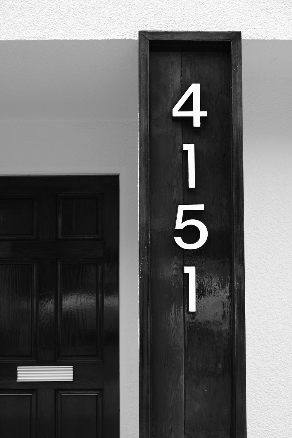 Black and white portrait photo of a house number on the side of a very expensive beach house. The metal numbers are positioned vertically inside a wooden box and read 4151.