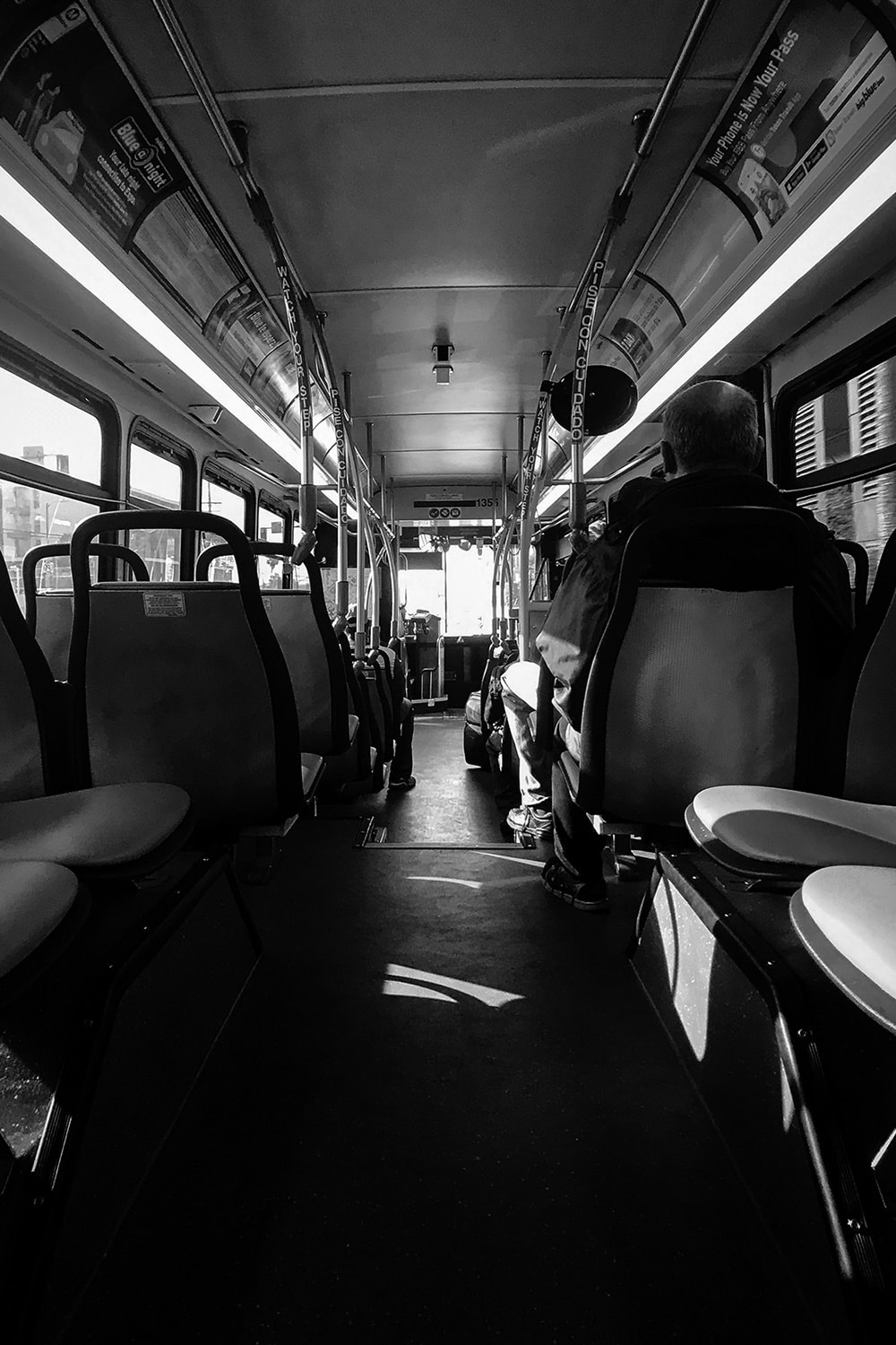 Black and white portrait photo of a man sitting on the bus. The photo is taken from the back of the bus looking forward so the interior lights, and walkway create leading lines towards the bus driver.