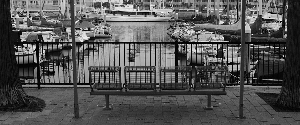 Black and white landscape photo of an empty metal wire bench looking into the rows of boats parked in one of the basins of Marina del Rey.