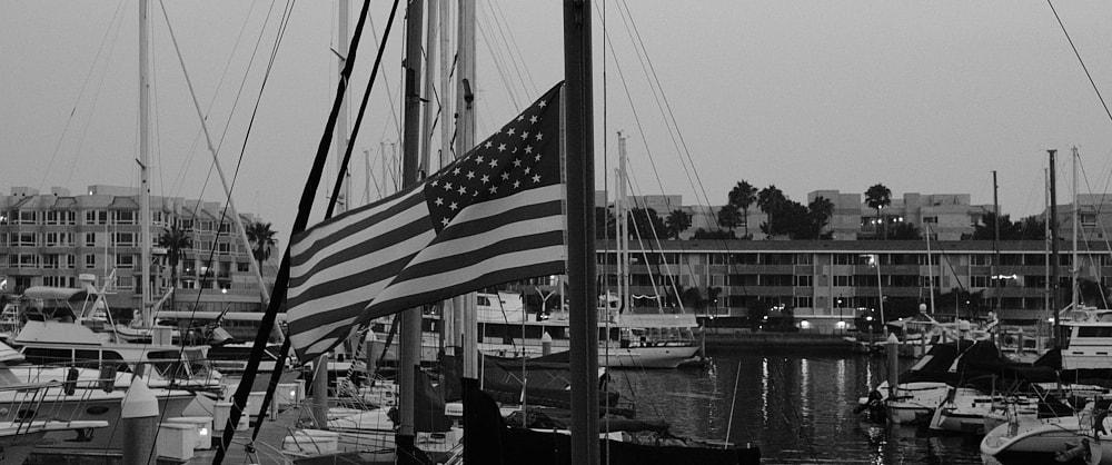 Black and white landscape photo of the American flag, waving in the breeze, attached to the mast of a sail boat.