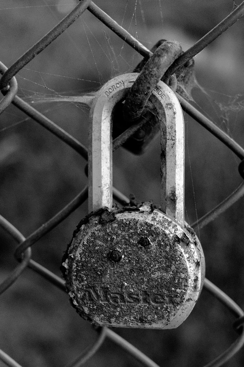 Black and white portrait photo of a rusty beaten up lock, locked to a chain link fence.
