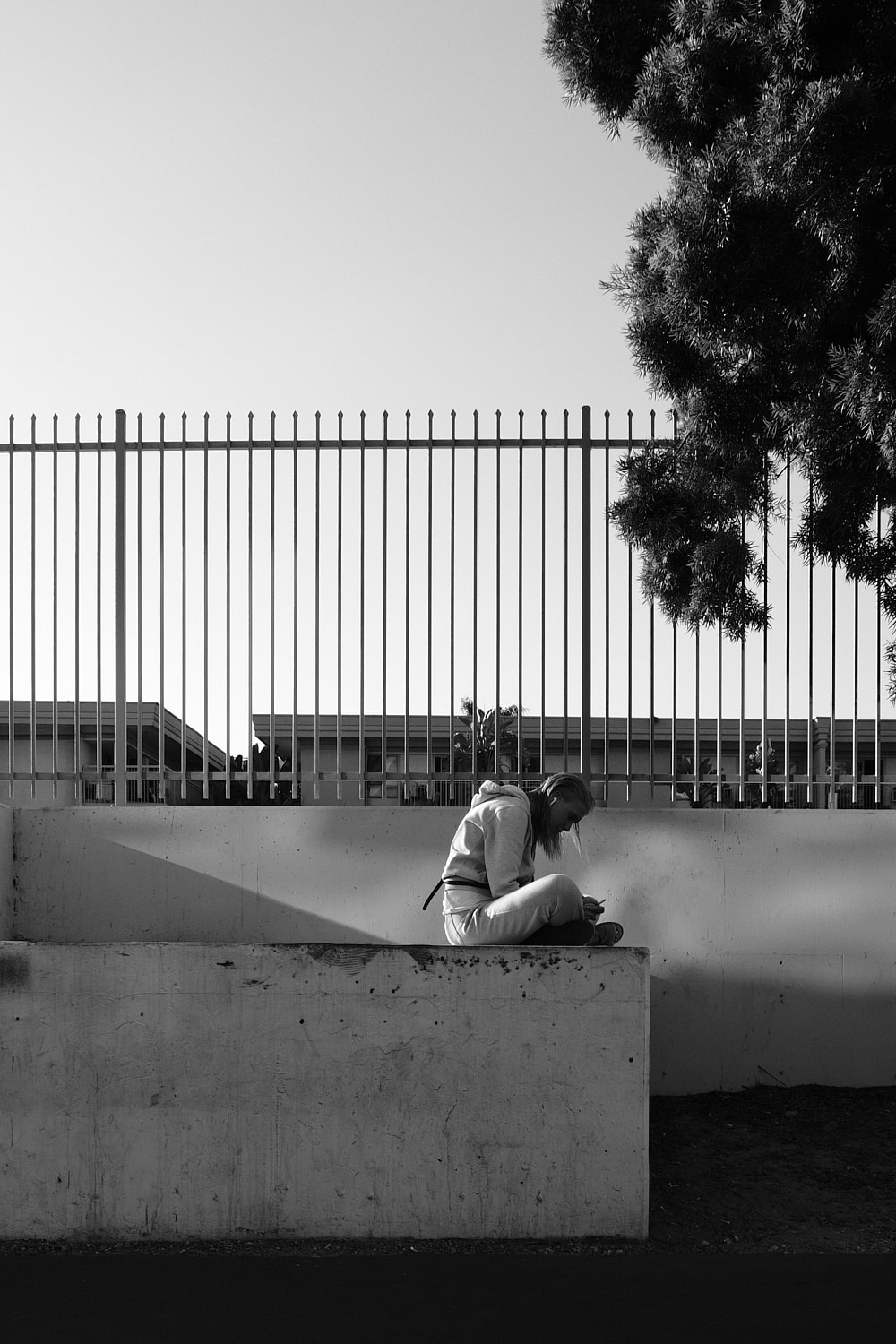 Black and white portrait photo of a woman sitting cross-legged in sweatpants and a hoodie on a white concrete wall. Her head is down as she looks at her phone. A tall metal fence with small spikes on each post is just behind the sitting woman.