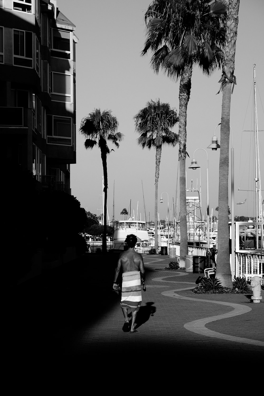 Black and white portrait photo of the back of a shirtless man wearing a towel around his waits walking away from you. He just steps into the sunlight from the shadow of the apartment building on the left.