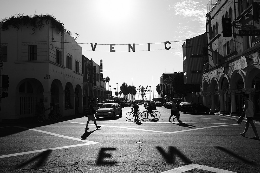 Black and white landscape photo of a crosswalk in Venice. The word V, E, N, I, C, E is spelled out in lights above the crosswalk. People and bikes move throughout the crosswalk.