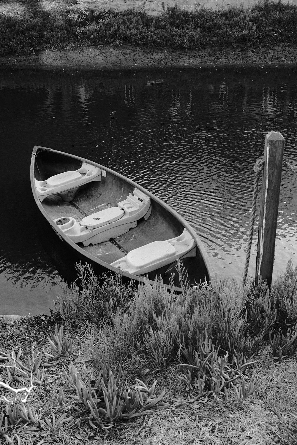 Black and white portrait photo of a canoe tied to a wooden post in the Venice canals.