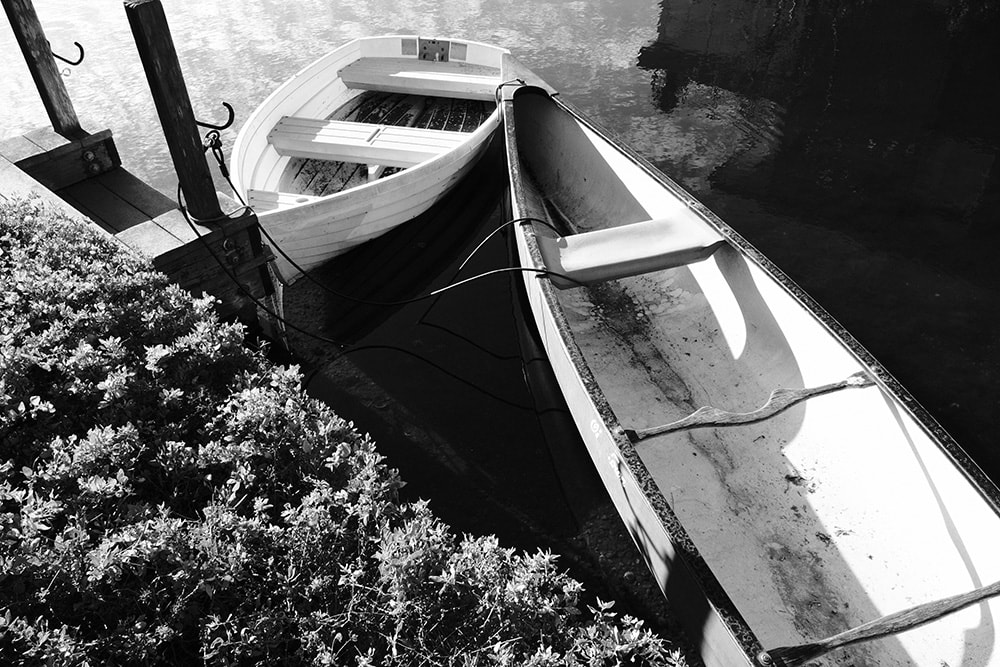 Black and white landscape photo of a rowboat and canoe tied to a dock in the Venice canals.