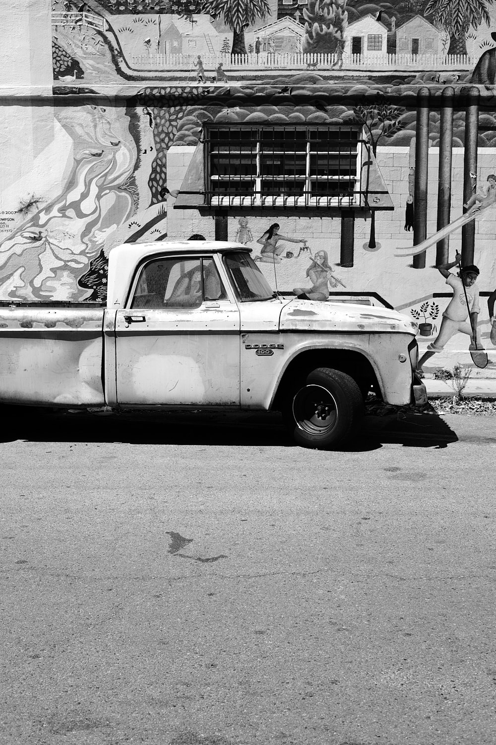 Black and white portrait photo of a 1960-70's beat up Dodge pickup truck parked in front of a wall mural with iron bars over a window.
